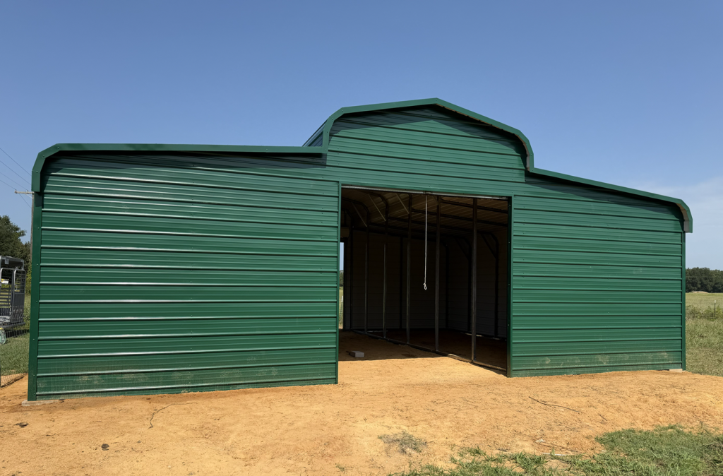 Green metal barn with open gable end and overhead door pull, on a gravel pad