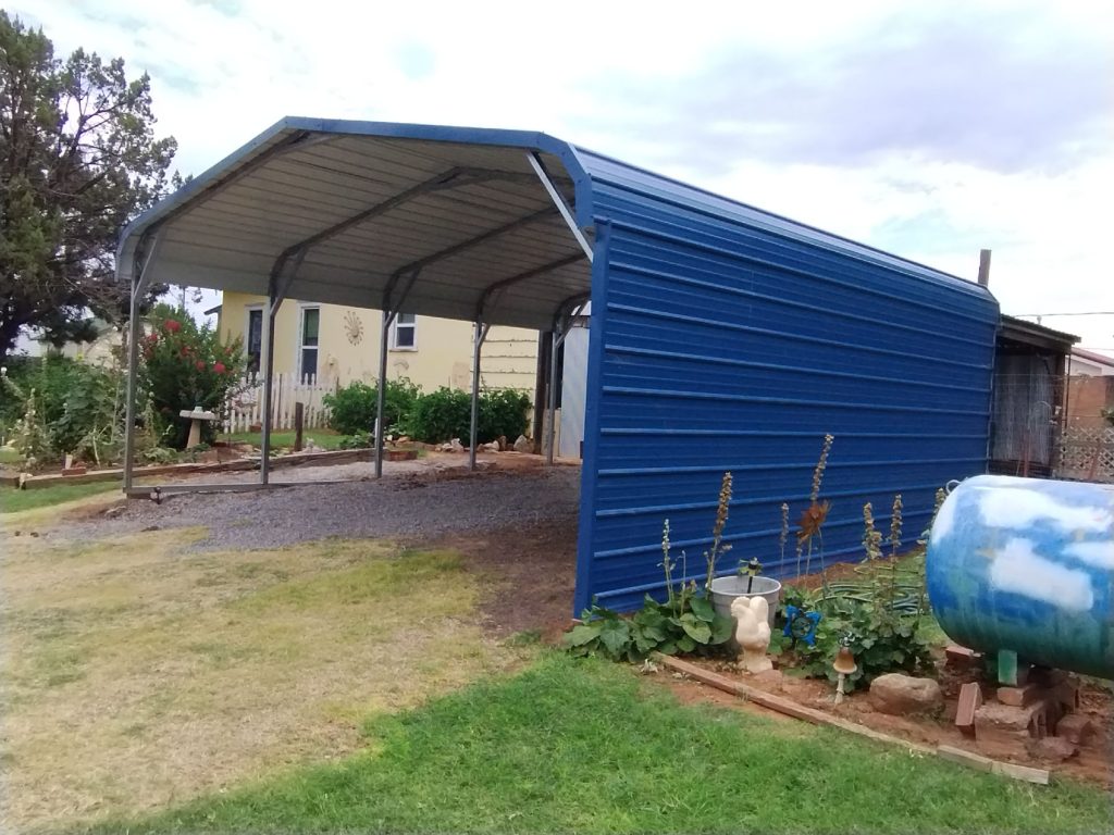 Blue metal carport-to-garage combo attached to a home, with garden beds