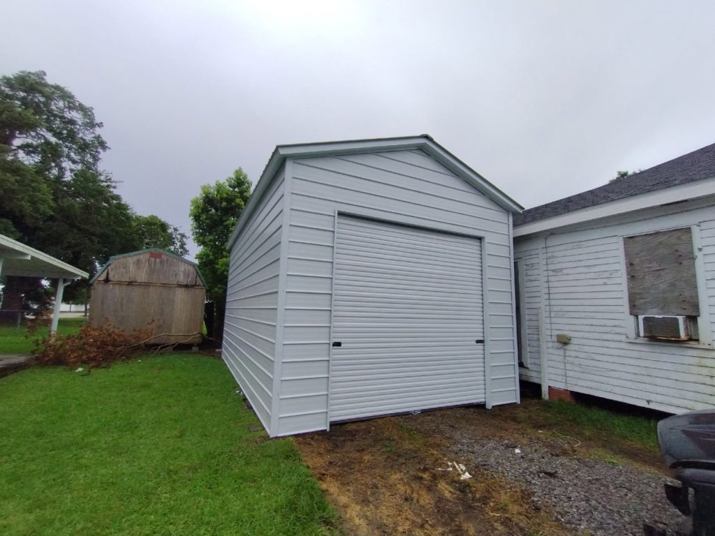 Gray metal storage shed with roll-up door beside an older wood-sided shed